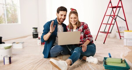 Cheerful Caucasian young married couple wife and husband sitting on floor in apartment near ladder, speaking on video call online using laptop computer. House repair concept. Videochatting on webcam