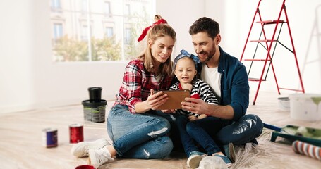 Happy Caucasian little adorable girl sitting with parents, tapping and browsing online on tablet,...