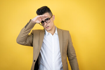 Young business man over isolated yellow background very happy and smiling looking far away with hand over head. searching concept.