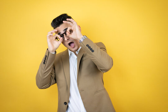 Young business man over isolated yellow background doing ok gesture shocked with smiling face, eye looking through fingers