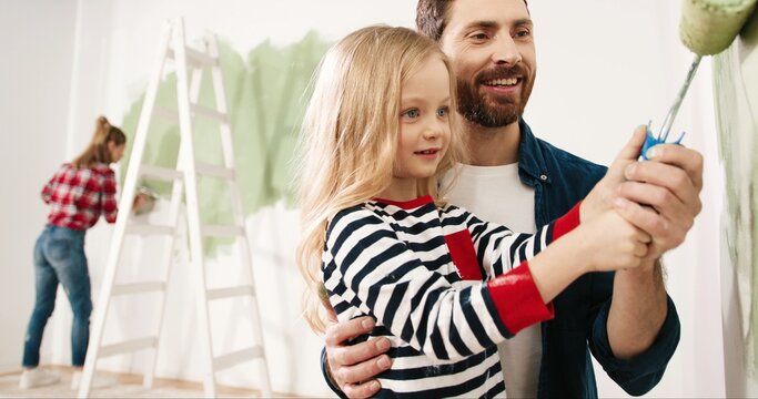 Portrait of cute family handsome happy father teaching little adorable kid girl daughter how to paint wall using paintbrush. Mother painting on background. Home renovation and repair concept