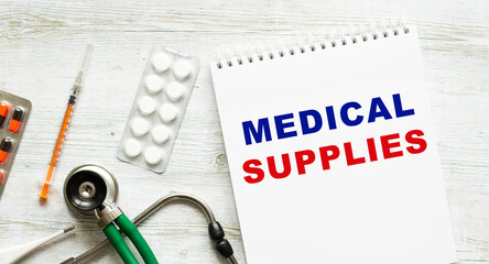 MEDICAL SUPLIES is written in a notebook on a white table next to pills and a stethoscope.