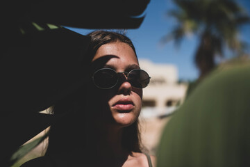 cute young teenage girl or woman posing with banana tree leaves dressed in summer hipster vintage white stylish clothes. Trendy girl posing. Funny and positive woman in sunglasses