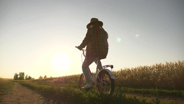 kid girl pedals on bicycle on rural road at sunset. Happy kid cyclist rides bicycle. Silhouette at sunset of girl in hat rides bicycle in field.back view.Happy child in countryside