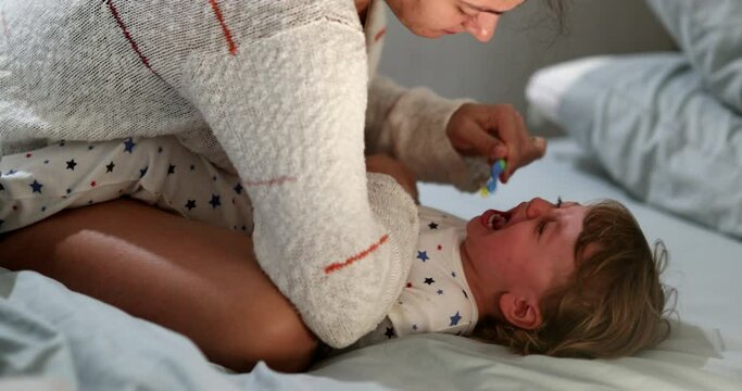 Candid mother brushing crying baby teeh. Upset infant boy not wanting to brush teeth