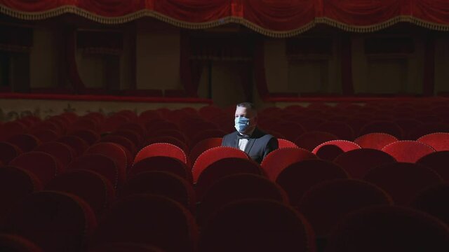 Rich man among rows of empty red chairs. Businessman in medical mask sitting in comfortable chairs alone on dark theater background. Pandemic in the world.