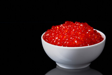 Red caviar in a white cup on a black background with a reflection.