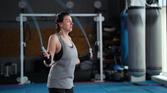 Fit woman with bandages on hands jumping on skipping rope in gym, equipment on blurred background. Side view athletic female warming up before boxing. Concept of sport