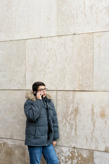 Young Caucasian student boy with glasses and modern coat talks to his friends with a smartphone leaning against a modern style wall. Vertical image with space for text
