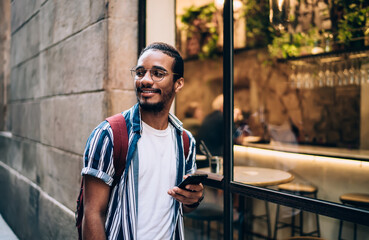 Handsome african american male standing on urban wall setting background using modern gadget connected to 4G internet, carefree dark skinned hipster guy smiling and holding mobile phone passing cafes