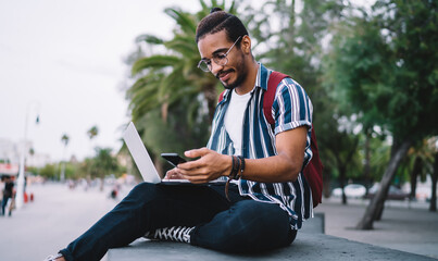 Fototapeta premium Young millennial dark skinned guy holding mobile phone reading information about online booking, hipster guys spending time time outdoors holding mobile phone working via laptop on freelance