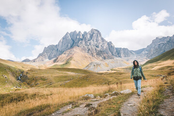Naklejka na ściany i meble Happy female caucasian woman walks down the path in Juta valley with scenic mountain landscape. KAzbegi travel destination