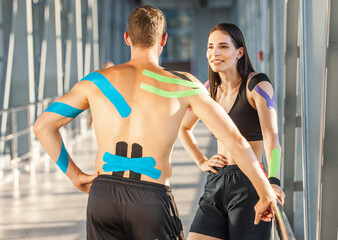Fit smiling brunette woman with hand on waist and man looking at smartphone and chatting. Young couple athletes posing indoors, colorful kinesiotaping on body, futuristic interior.