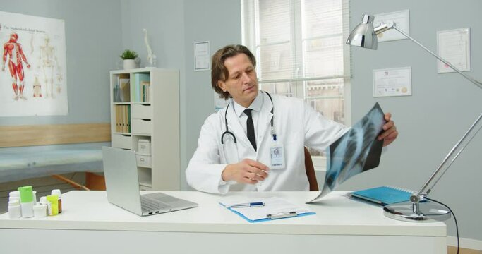 Portrait of busy Caucasian middle-aged man doctor in medical coat sitting at esk in hospital room, working on laptop, looking at X-ray scan results. coronavirus pandemic. treatment concept