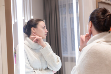 Obraz na płótnie Canvas Young beautiful woman in a sweater in a beauty salon looks in the mirror, touches her face, thinks about the upcoming procedures, considers herself