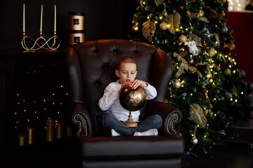 The boy in the armchair in a dark home room with Christmas decor. Boy looking at globe.