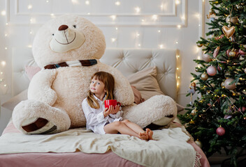 Beautiful little girl in a Christmas room drinking coffee in a bed with large Teddy bear plush toy.