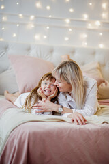 Mom and her little daughter have fun and play sitting on the bed in a bright bedroom with Christmas decor.