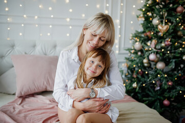 Happy mother and daughter cuddle while lying on the bed in front of a Christmas tree decorated with a garland.
