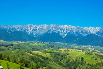 Fototapeta na wymiar Stunning alpine landscape with green fields and high snowy Piatra Craiului mountains near Brasov. Panoramic view of mountain farms with houses. Bran, Transylvania, Romania, Europe. 