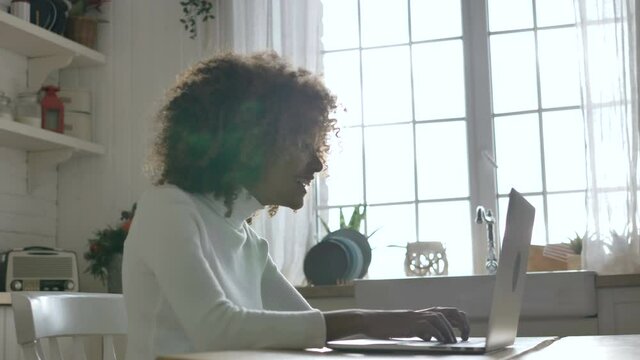 Cheerful African-American woman with curly hair enjoys the good news, typing on a gray notebook, sitting in a modern kitchen close-up slow motion