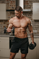 Obraz premium A muscular man with a beard is training his biceps with dumbbells in his apartment. A bodybuilder with tattoos on his arms is warming up during a workout at home. Athlete with a naked torso in shorts