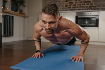 A muscular man with a beard is doing pushups on a blue yoga mat in his apartment in the evening. An athletic guy with tattoos on his forearms is doing a chest and triceps workout at home.