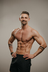 Fototapeta premium A smiling muscular man with a beard is posing. The athletic guy is demonstrating his sporty physique. A bodybuilder with tattoos on his forearms is standing with his hands on his hips