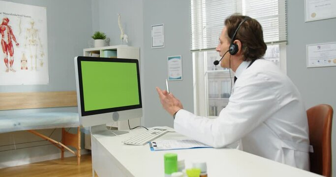 Side view. Portrait of Caucasian male physician in headset sitting at desk in clinic cabinet, videochatting on video call though webcam on computer looking at green screen. Monitor with chroma key