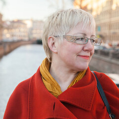 Mature blonde shorthaired woman  in red coat, golden glasses and yelllow glowes on Moika river embankment