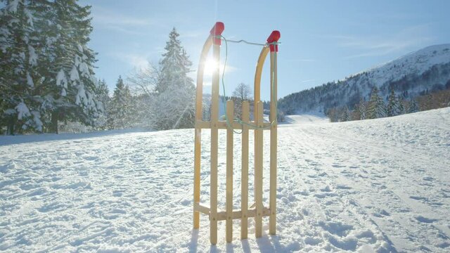 LENS FLARE: Wooden sled is illuminated by the golden winter sun rays while standing upright in the middle of a snowy meadow in Kranjska Gora. Sleigh is set aside during a fun day in wintry mountains