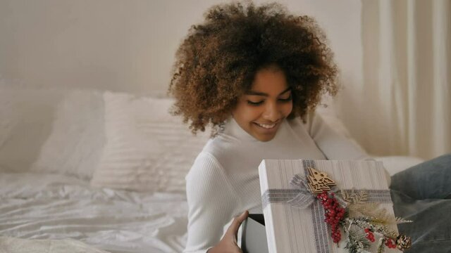 Lady with short kinky hair and dark skin sits holding present box against designer white Christmas tree closeup slow motion