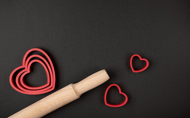 Red heart-shaped baking tins for cookies and wooden rolling pin on the dark (black) background....