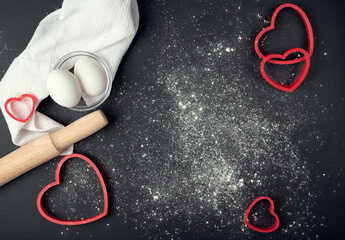 Red heart-shaped baking tins for cookies and wooden rolling pin with scattered flour on the dark (black) background. Flat lay, top view, space for text. Valentine's day concept.