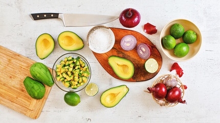 Avocado halves, pieces, limes and onions, cup with salt - basic guacamole ingredients and chef knife on white working board, flat lay photo