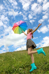 Child a girl with a great mood jumps in the summer in a field with a rainbow umbrella on which water pours from the sky.