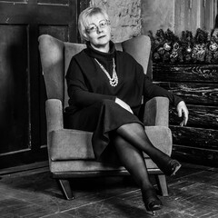 Mature woman in glasses with short blond hair sits in a leather armchair in a loft