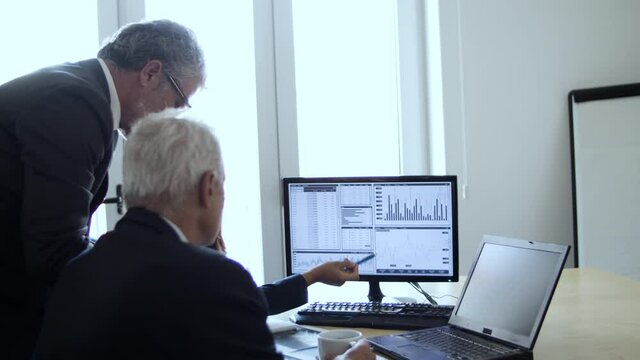 Busy traders analyzing stock market statistics, looking and pointing at financial diagrams on pc monitor, talking and writing notes. Business and trading concept