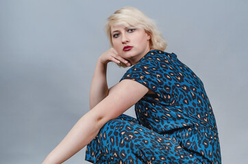 Blonde woman in spotted dress squats, close-up portrait side view, isolate on gray background