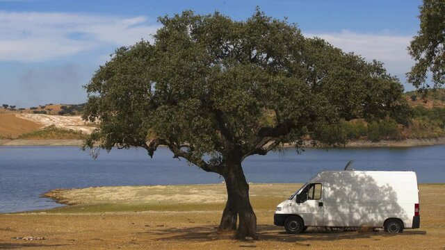 Camper van on an Alentejo dam landscape with the lake behind and under the shade of a tree, in Portugal
