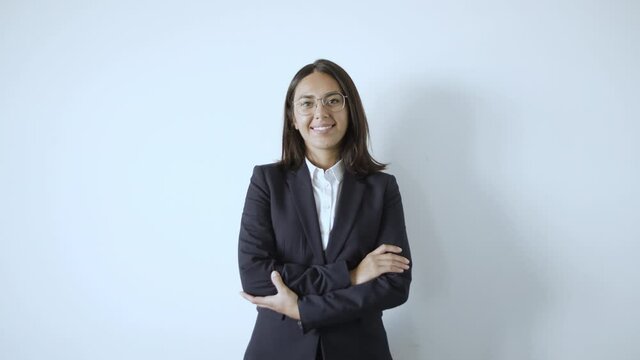 Happy successful young businesswoman wearing suit, posing at white wall, standing and crossing arms, looking at camera and smiling. Front view. Business portrait concept