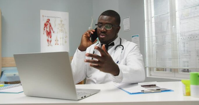 Portrait of young serious African American general practitioner physician sitting at table in cabinet in medical center typing on laptop and talking on cellphone. Healthcare. clinic concept