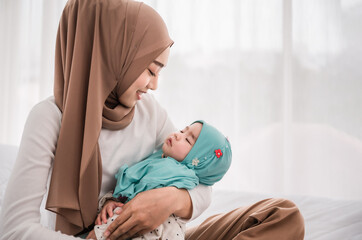 Happy muslim mother holding adorable little baby daughter wearing hijab in her arms on white bed in bedroom.