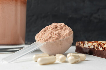 Scoop of whey or soy protein isolate, chocolate protein bar, white capsules of amino acids and creatine, bodybuilding food supplements on white table and dark background with copy-space