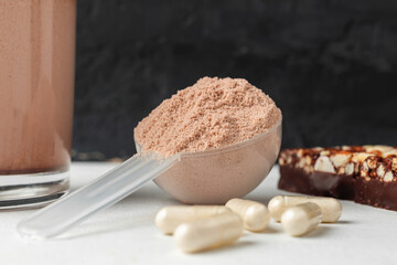 Scoop of whey or soy protein isolate, chocolate protein bar, white capsules of amino acids and creatine, bodybuilding food supplements on white table and dark background with copy-space