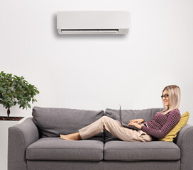Woman working on a laptop and laying on a sofa at home under an air conditioning unit
