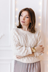 Naklejka premium beautiful young woman in a white sweater, skirt and high heels on a background of white wall and christmas garlands