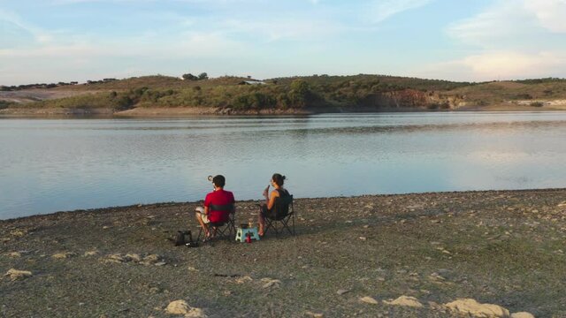Caucasian couple sit on chairs on a lake drinking beer and seeing the landscape in Alentejo, Portugal