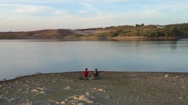 Caucasian couple sit on chairs on a lake drinking beer and seeing the landscape in Alentejo, Portugal