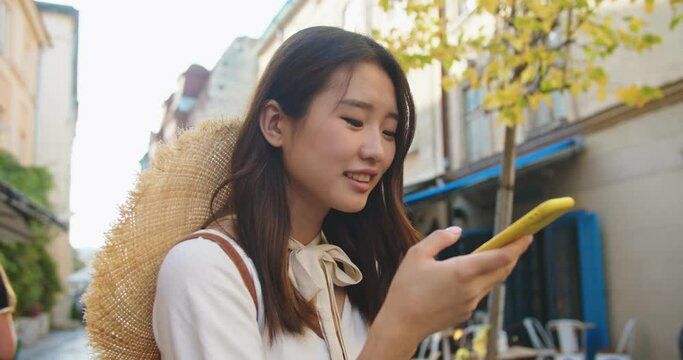 Close up portrait of joyful young female tourist with hat standing on street and recording voice message on speakerphone. Happy smiling Asian woman using virtual assistant app on cellphone in city.